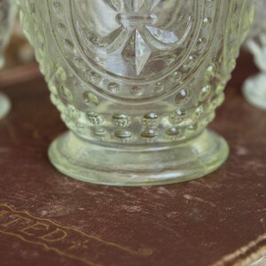 May include: Close-up of three clear, vintage-style glass goblets. The goblets have a detailed, embossed design with a starburst pattern and a rounded base. The goblets are on a brown surface.