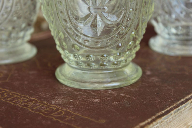 May include: Close-up of three clear, vintage-style glass goblets. The goblets have a detailed, embossed design with a starburst pattern and a rounded base. The goblets are on a brown surface.