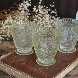 May include: Three clear, vintage-style glass votive candle holders with ornate embossed patterns. The candle holders are on a worn, antique book with the title "HIGHLY ILLUSTRATED". White dried flowers and a decorative snow globe are in the background.