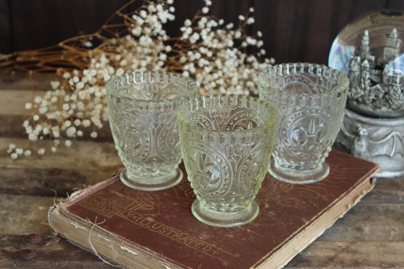 May include: Three clear, vintage-style glass votive candle holders with ornate embossed patterns. The candle holders are on a worn, antique book with the title "HIGHLY ILLUSTRATED". White dried flowers and a decorative snow globe are in the background.