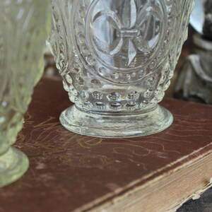 May include: Clear glass goblets with ornate detailing, including a fleur-de-lis design, sit on a vintage book with a worn, brown cover. The goblets have a textured surface and are partially in focus.