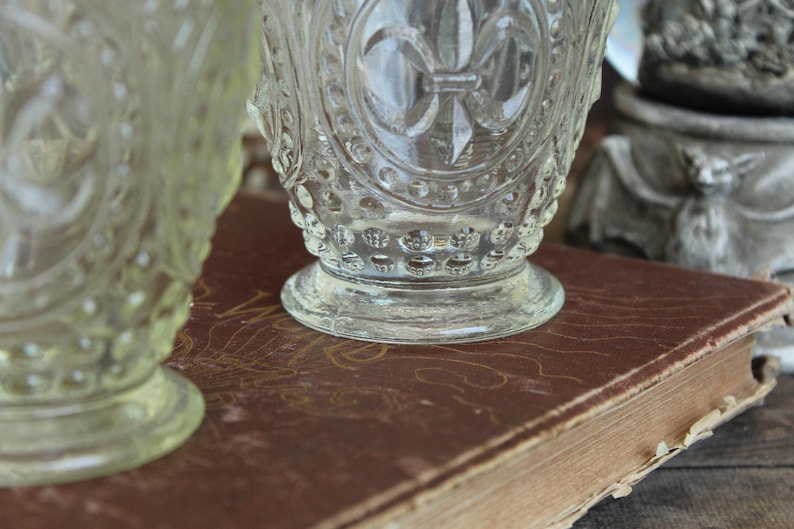 May include: Clear glass goblets with ornate detailing, including a fleur-de-lis design, sit on a vintage book with a worn, brown cover. The goblets have a textured surface and are partially in focus.