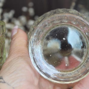 May include: Clear glass votive candle holder with a detailed, textured design. The bottom of the votive is visible, showing a rounded shape. The glass has a slightly yellow tint. The votive is held in a hand.