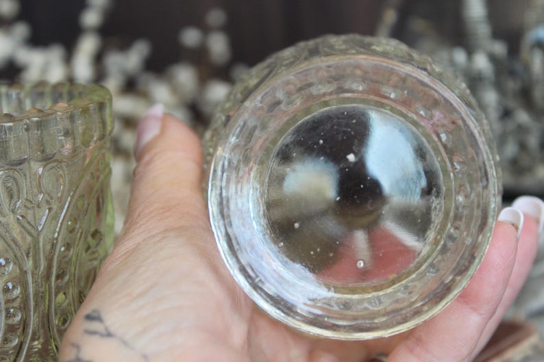 May include: Clear glass votive candle holder with a detailed, textured design. The bottom of the votive is visible, showing a rounded shape. The glass has a slightly yellow tint. The votive is held in a hand.