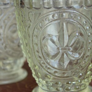 May include: Clear glass goblets with ornate embossed designs. The goblets feature a fleur-de-lis emblem and other decorative patterns. The glass has a textured surface and a rounded base. The goblets are likely vintage or antique.
