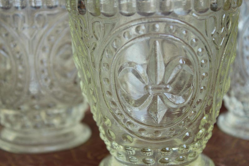 May include: Clear glass goblets with ornate embossed designs. The goblets feature a fleur-de-lis emblem and other decorative patterns. The glass has a textured surface and a rounded base. The goblets are likely vintage or antique.