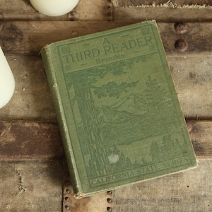 May include: A vintage green hardcover book titled A Third Reader by Brooks from the California State Series. The cover features a dark green embossed illustration of a mountain landscape with trees and a lake, alongside two white pillar candles.
