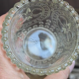 May include: Clear glass votive candle holder with ornate etched floral and scroll designs. The rim features a beaded edge. The holder is held in a hand, with the interior reflecting light.