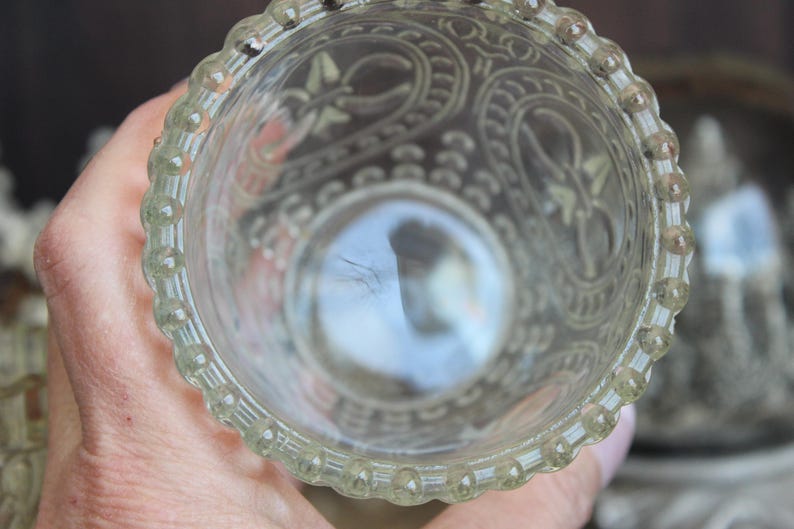 May include: Clear glass votive candle holder with ornate etched floral and scroll designs. The rim features a beaded edge. The holder is held in a hand, with the interior reflecting light.