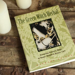 May include: A book titled "The Green Witch Herbal" with a green cover and a central image. The book is on a rustic wooden surface, accompanied by two white candles and small white flowers. The book's subtitle is "Restoring Nature's Magic in Home, Health & Beauty Care."