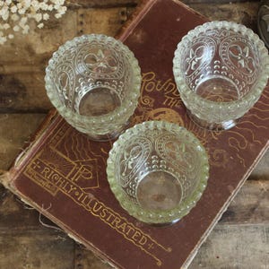 May include: Three clear glass votive candle holders with ornate floral designs are arranged on a vintage book. The book has gold lettering that reads "RICHLY ILLUSTRATED". The candle holders are on a wooden surface.