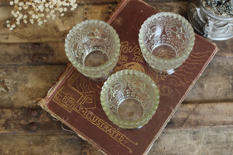 May include: Three clear glass votive candle holders with ornate floral designs are arranged on a vintage book. The book has gold lettering that reads "RICHLY ILLUSTRATED". The candle holders are on a wooden surface.