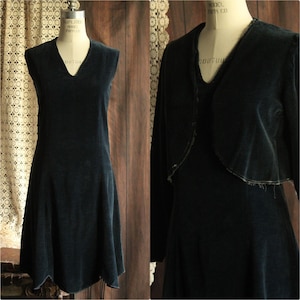 May include: A dark teal velvet dress with a V-neck and a flared skirt is displayed on a mannequin. A matching bolero jacket is draped over the dress. The dress and jacket are vintage and have a soft, textured appearance.