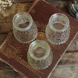May include: Three clear, vintage glass inkwells with ornate, embossed patterns. The inkwells are arranged on a worn, antique book with gold lettering. The book is resting on a rustic wooden surface, creating a vintage aesthetic.