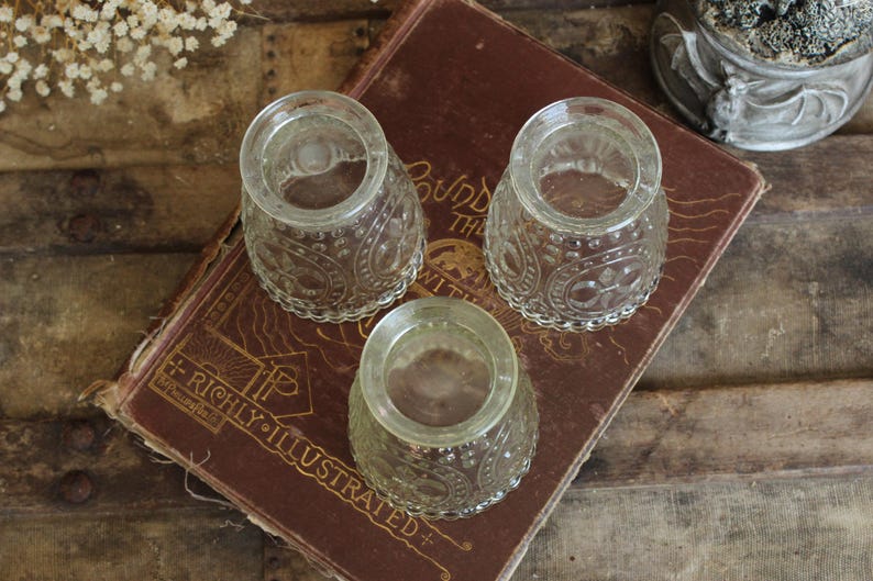 May include: Three clear, vintage glass inkwells with ornate, embossed patterns. The inkwells are arranged on a worn, antique book with gold lettering. The book is resting on a rustic wooden surface, creating a vintage aesthetic.