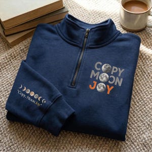 May include: Navy blue quarter-zip sweatshirt with the text "COPY MOON JOY" and moon phase graphics. The sweatshirt has a silver zipper and is made of a soft material. The text and graphics are in silver and orange.