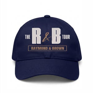 May include: Navy blue baseball cap with the text "THE R&B TOUR" in white and brown. The text "RAYMOND & BROWN" is printed below the main text. The cap has a curved brim and a button on top.