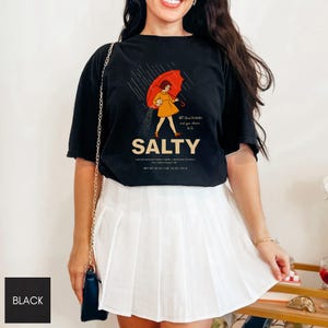 May include: Black t-shirt with the word "SALTY" in a bold font. The shirt features a graphic of a person under a red umbrella in the rain. The text "All these flavors and you choose to be" is above the word "SALTY".