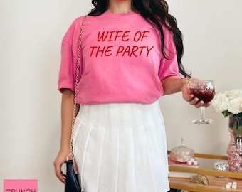 May include: A pink t-shirt with the text "WIFE OF THE PARTY" in red. The shirt is paired with a white pleated skirt. The image also shows a glass of red wine and a small black purse with a gold chain.