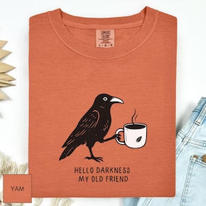 May include: A burnt orange Comfort Colors t-shirt featuring a black crow illustration holding a white coffee mug. The text "HELLO DARKNESS MY OLD FRIEND" is below the crow. The shirt has a crew neck and short sleeves.