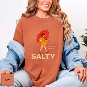 May include: A rust-colored t-shirt with a graphic of a person holding a red umbrella in the rain. The word "SALTY" is below the illustration, with text that reads "All these flavors and you choose to be." The shirt's net weight is 26 oz.