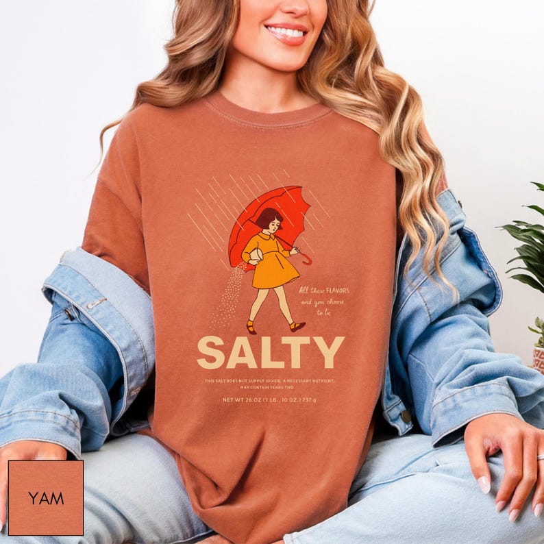 May include: A rust-colored t-shirt with a graphic of a person holding a red umbrella in the rain. The word "SALTY" is below the illustration, with text that reads "All these flavors and you choose to be." The shirt's net weight is 26 oz.