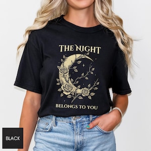 May include: Black t-shirt with the text "THE NIGHT BELONGS TO YOU" in gold. The design features a crescent moon with roses and branches. The shirt is a crew neck. The person is wearing blue jeans.