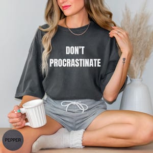 May include: A dark gray t-shirt with the words "DON'T PROCRASTINATE" in white block letters. The person is wearing gray shorts and white socks, holding a white mug. The shirt is a casual style.