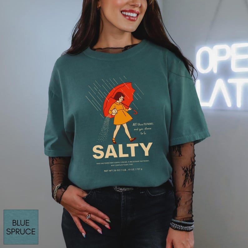 May include: Blue spruce-colored t-shirt with a graphic of a person under a red umbrella, with the word "SALTY" below. The shirt also has the text "All the flavors and you choose to be." The shirt's net weight is 26 oz.