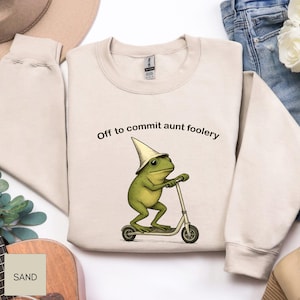 May include: A sand-coloured sweatshirt with the phrase "Off to commit aunt foolery." A green frog wearing a white hat and riding a scooter is the graphic. The sweatshirt is made of a soft material.