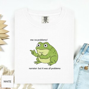 Peut inclure: T-shirt blanc avec une grenouille verte dessinée, pointant du doigt et le texte "me: no problemo!" et "narrator: but it was all problemo". Le t-shirt a une étiquette Comfort Colors.