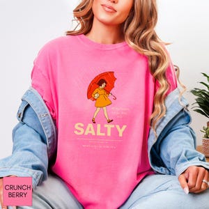 May include: A pink sweatshirt with a graphic of a person holding an orange umbrella in the rain. The word "SALTY" is printed in large, yellow letters below the illustration. The sweatshirt also has text at the bottom and a pink label in the bottom left corner.