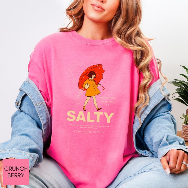 May include: A pink sweatshirt with a graphic of a person holding an orange umbrella in the rain. The word "SALTY" is printed in large, yellow letters below the illustration. The sweatshirt also has text at the bottom and a pink label in the bottom left corner.