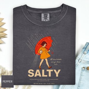 May include: Dark gray Comfort Colors t-shirt with a graphic of a person under a red umbrella, with the word "SALTY" below. The graphic includes the text "All these flavors and you choose to be." The shirt's net weight is 26 oz (1 lb., 10 oz.) 737 g.