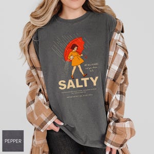 May include: Dark gray t-shirt with a graphic of a person under a red umbrella, with the word "SALTY" below. The shirt also has the text "All the flavors and you choose to be". The shirt is made of a soft material.