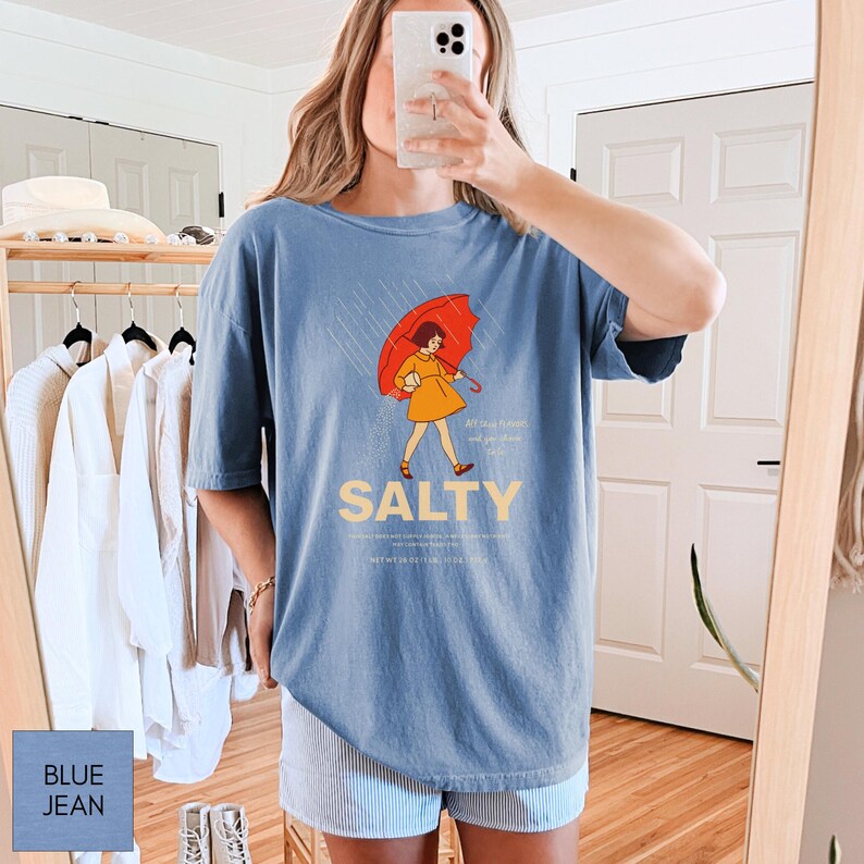May include: Blue jean t-shirt with a graphic of a person under a red umbrella in the rain. The word "SALTY" is printed in a light tan color below the graphic. The shirt is a light blue color.