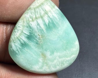 May include: A teardrop-shaped gemstone with a light turquoise color and white veining. The stone has a polished surface and is held in a hand. The gemstone's pattern resembles ocean waves.