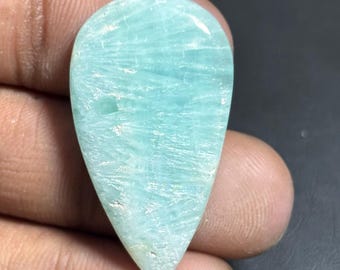 May include: A polished, teardrop-shaped gemstone with a light blue-green hue. The stone has a textured surface with white veining, and is held in a hand against a dark background. The gemstone is likely used for jewelry or decorative purposes.