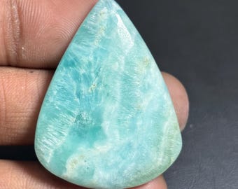 May include: A polished, teardrop-shaped gemstone with a light blue and white color pattern. The stone has a smooth surface and a translucent appearance, with visible internal veining. The gemstone is held in a hand against a dark background.
