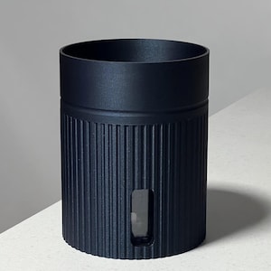 May include: A matte black cylindrical container featuring a vertical ribbed texture along the exterior body. The top section is smooth and solid, while a small, rectangular transparent window is integrated into the lower ribbed portion of the vessel.