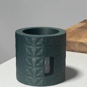May include: A dark teal cylindrical container with a geometric pattern and a rectangular cutout. The container has a wide opening at the top and sits on a white surface. The object appears to be 3D printed.
