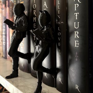 Può includere: Ferma libri neri a forma di figure che leggono, appoggiate a una fila di libri scuri. Una figura ha un design a orecchie di gatto. I libri hanno testo bianco sui dorsi. I fermalibri sono su una mensola bianca.