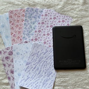 Può includere: Un e-reader nero con il logo Amazon è accanto a un ventaglio di carte decorate. Le carte presentano disegni floreali e botanici nei toni del rosa, viola e blu. I motivi includono fiocchi, strisce e rametti di lavanda.