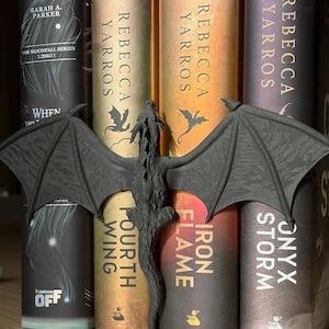 Può includere: Una statuina di drago nero con le ali spiegate è appoggiata davanti a una pila di libri. I libri hanno copertine dorate e scure con titoli come "Fourth Wing" e "Iron Flame" di Rebecca Yarros. I libri sono su una superficie di legno.