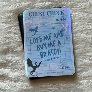 Può includere: Un conto del ristorante olografico con la scritta "LOVE ME AND BUY ME A DRAGON". Il conto ha uno sfondo blu con sagome di draghi e il numero del conto 037594. Il conto è sopra un oggetto nero.
