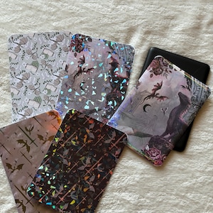 Può includere: Un assortimento di custodie decorative per e-reader con vari design. Una custodia presenta un motivo floreale e di pipistrelli, mentre altre mostrano effetti olografici con illustrazioni gotiche. È visibile anche un e-reader nero.