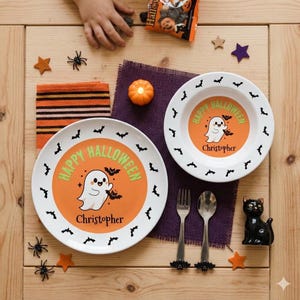 May include: A white dinner plate and a matching bowl, both featuring an orange center with a cartoon ghost, bats, and the text Happy Halloween Christopher. The rim is white with black bat silhouettes. A silver fork and spoon are included.