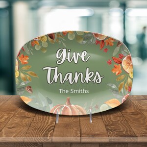 May include: Oval decorative plate with a green background and the words "Give Thanks" in white script. The plate is adorned with fall-themed illustrations of pumpkins, leaves, and oranges. The name "The Smiths" is printed below the text.