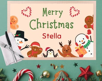 May include: A festive Christmas placemat with the words "Merry Christmas Stella" in green and red. The placemat features gingerbread men, a snowman, a penguin, and a reindeer. A fork, knife, and napkin are on the left.
