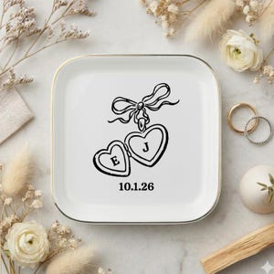 May include: A square white ceramic trinket dish with a gold-toned rim. The center features a black line drawing of a heart-shaped locket tied with a bow, with the letters E and J inside the hearts and the date 10.1.26 printed below in black.